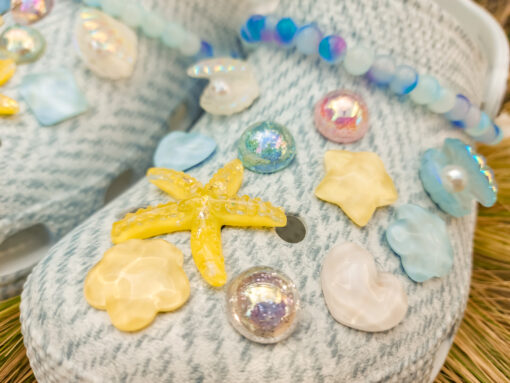 A 24-piece beach-themed charm and chain set on a pair of light gray woven-texture shoes. The set includes sea-inspired charms like starfish, shells, pearls, and iridescent accents with two beaded chains draped across the tops, evoking an oceanic look