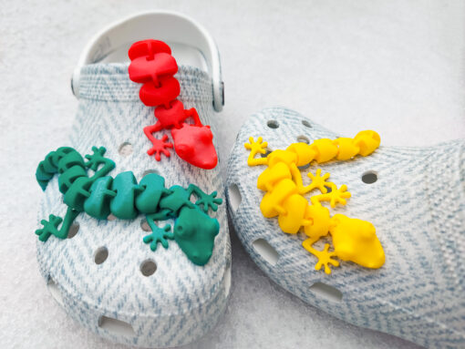 Three colorful 3D printed flexi lizard charms in red, green, and yellow, clipped onto blue-patterned shoes. The articulated lizards appear lifelike with detailed feet and movable segments, set against a snowy white backdrop.