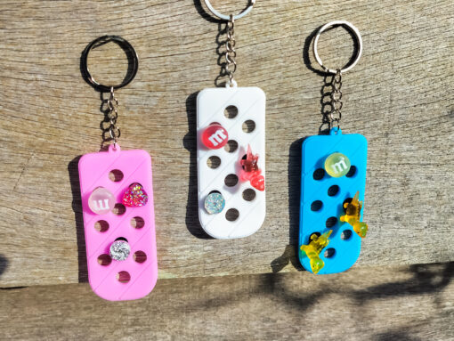 Three PVC keychains in pink, white, and blue with holes designed to hold shoe charms. Each keychain displays a unique set of charms, including candy-themed and colorful glitter charms, showcasing a playful and customizable accessory for charm lovers.
